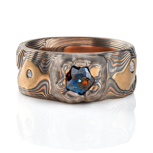 Large men's style mokume gane patterned ring, made to resemble rock formations and mountains, with a large dark blue and orange stone set into the ring in a raised area, intended to look like it's being excavated from the earth.